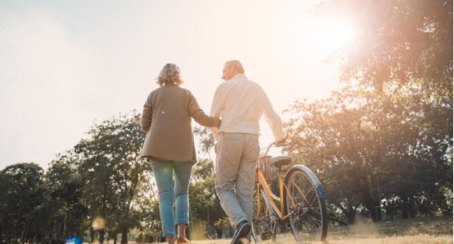 An older couple walking through a park as one pushes a bike.